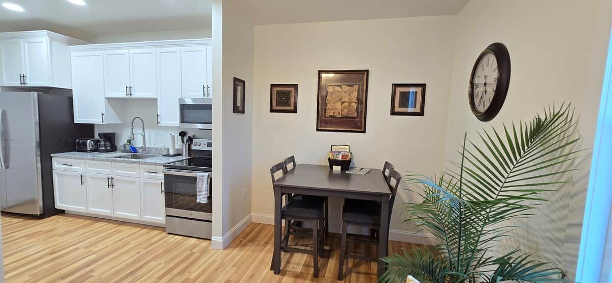 A modern kitchen with white cabinets, a microwave, and a sink. There is a dining table with chairs and a clock on the wall.