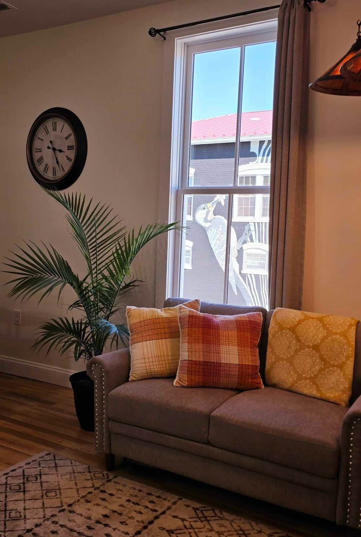 Una sala de estar con un sofá, una planta en maceta, un reloj de pared y una ventana con vistas a un edificio.