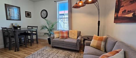 A living room with a sofa, a dining table with chairs, a potted plant, a clock on the wall, and a window with a view.