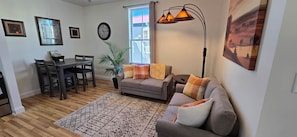 A living room with a sofa, a dining table, chairs, a lamp, and a window with curtains.