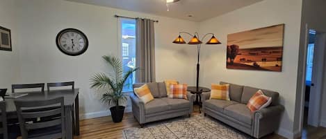 A living room with a dining table, two sofas, a floor lamp, a clock, and a window with curtains.