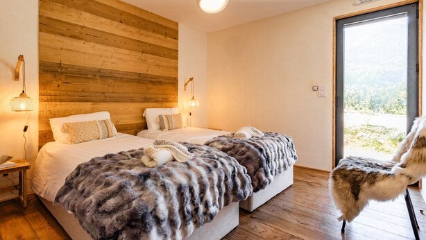 A bedroom with two beds, wooden paneling on the wall, and a view through a glass door.