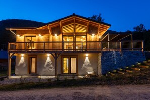A two-story wooden cabin with a balcony, large windows, and a stone wall.