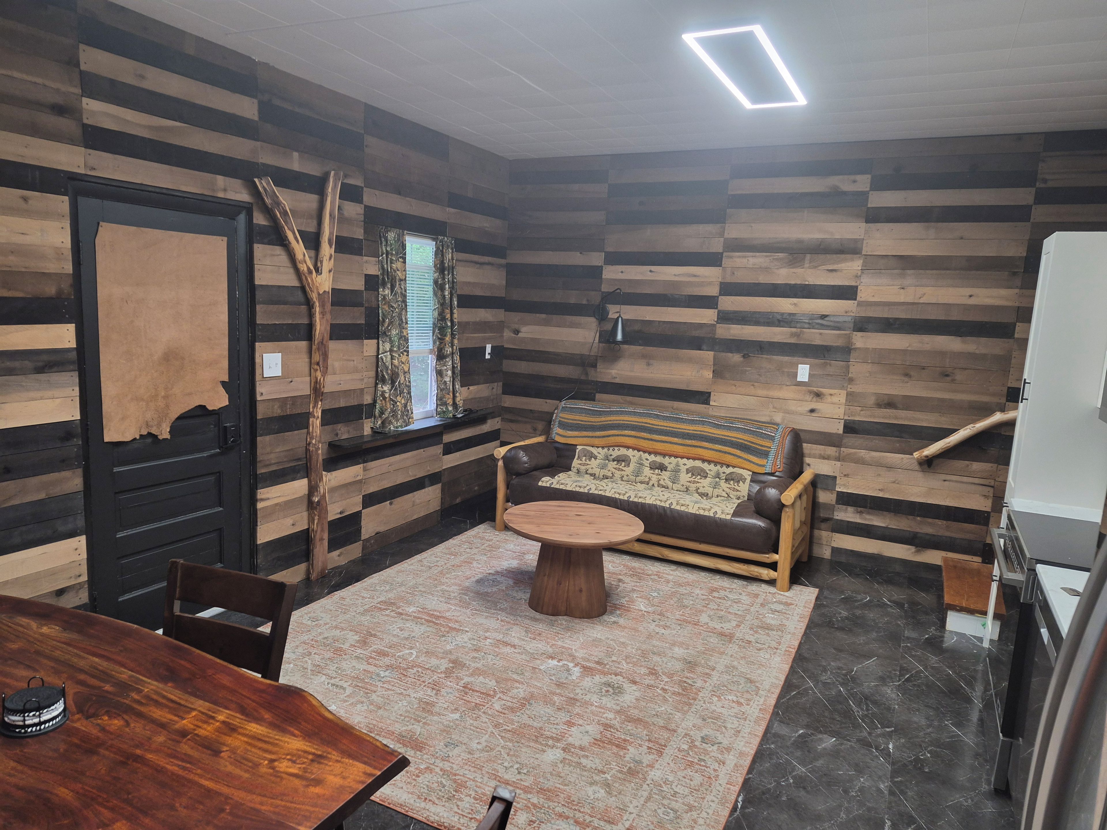 Una habitación con pared de madera, un sofá, una mesa redonda, una silla, una ventana con cortinas y una puerta.