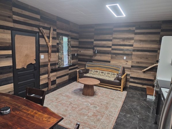 A room with a wooden wall, a sofa, a round table, a chair, a window with curtains, and a door.