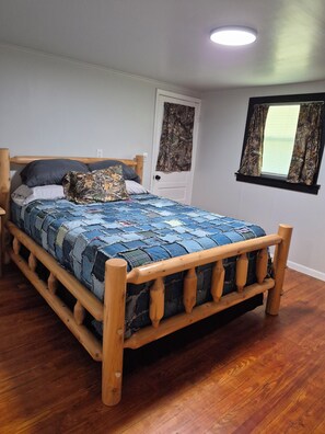 A wooden bed with a quilt, a door with a camouflage curtain, and a window with blinds.