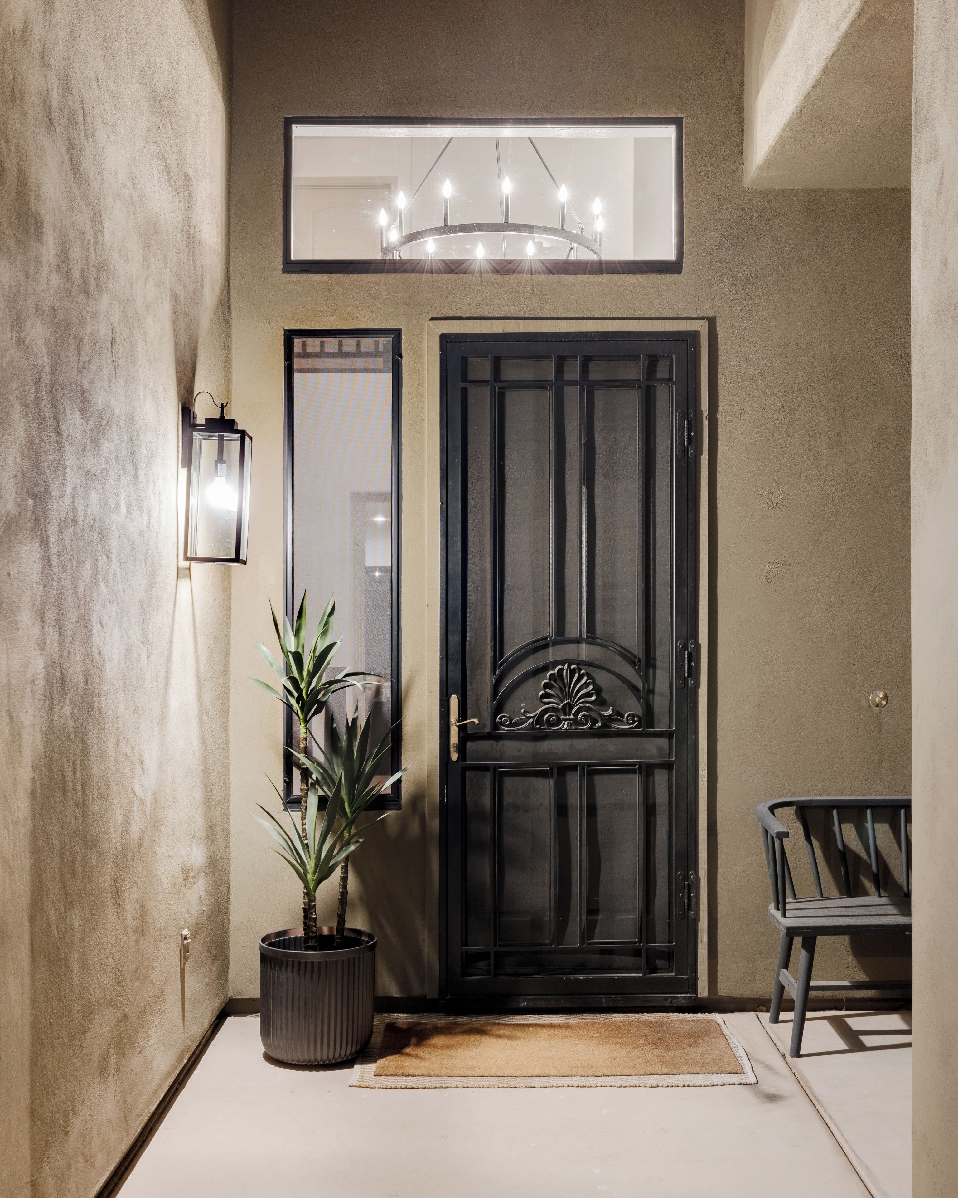 Una porta negra amb un panell decoratiu, una planta en test, un llum fixada a la paret i una cadira.