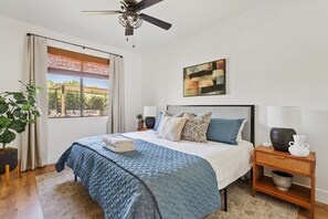 Ein Schlafzimmer mit einem Bett, einem Deckenventilator, einem Fenster mit Vorhängen, einer Pflanze, einem Gemälde und einem Nachttisch mit Lampe und Vase.