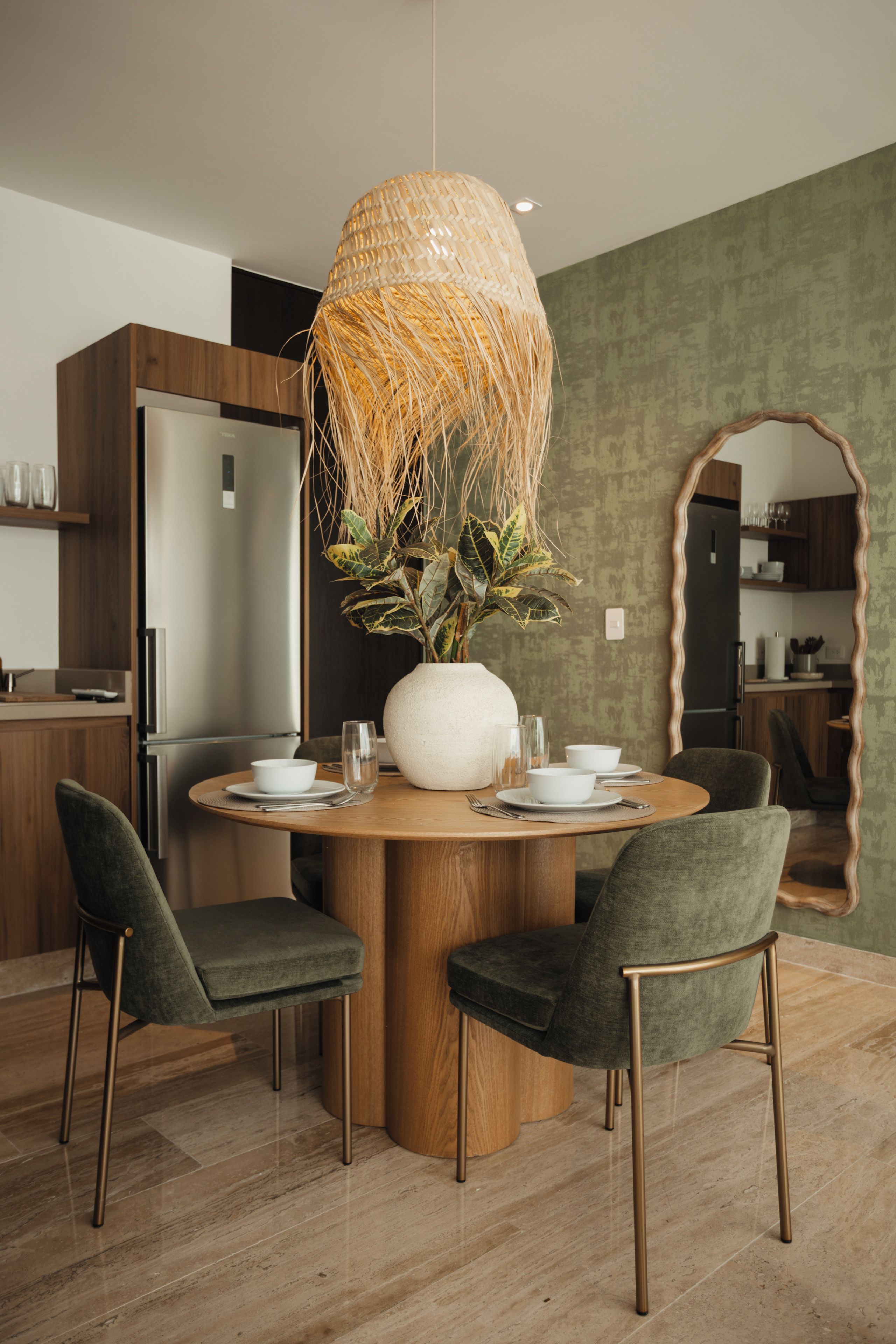 Un espace repas moderne avec une table ronde en bois, deux chaises, un grand luminaire en fibres tressées et un mur vert à texture.