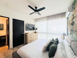 Ein Schlafzimmer mit einem Bett, einem Deckenventilator, einem Fernseher, einer Kommode und einem Fenster mit durchsichtigen Vorhängen.