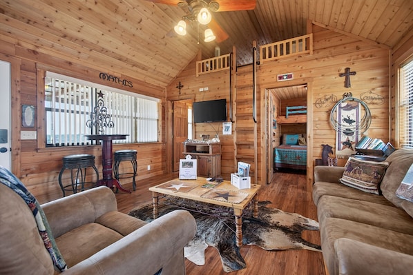 A cozy log cabin interior with a dining area, a television, and a living room furnished with sofas and a coffee table. - New! F'burg w/ Epic Views, Huge Covered Porch! (Fredericksburg)