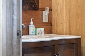 A bathroom with a wooden wall, a white sink, and toiletries including a soap dispenser, Dove body wash, and a razor. - New! F'burg w/ Epic Views, Huge Covered Porch! (Fredericksburg)