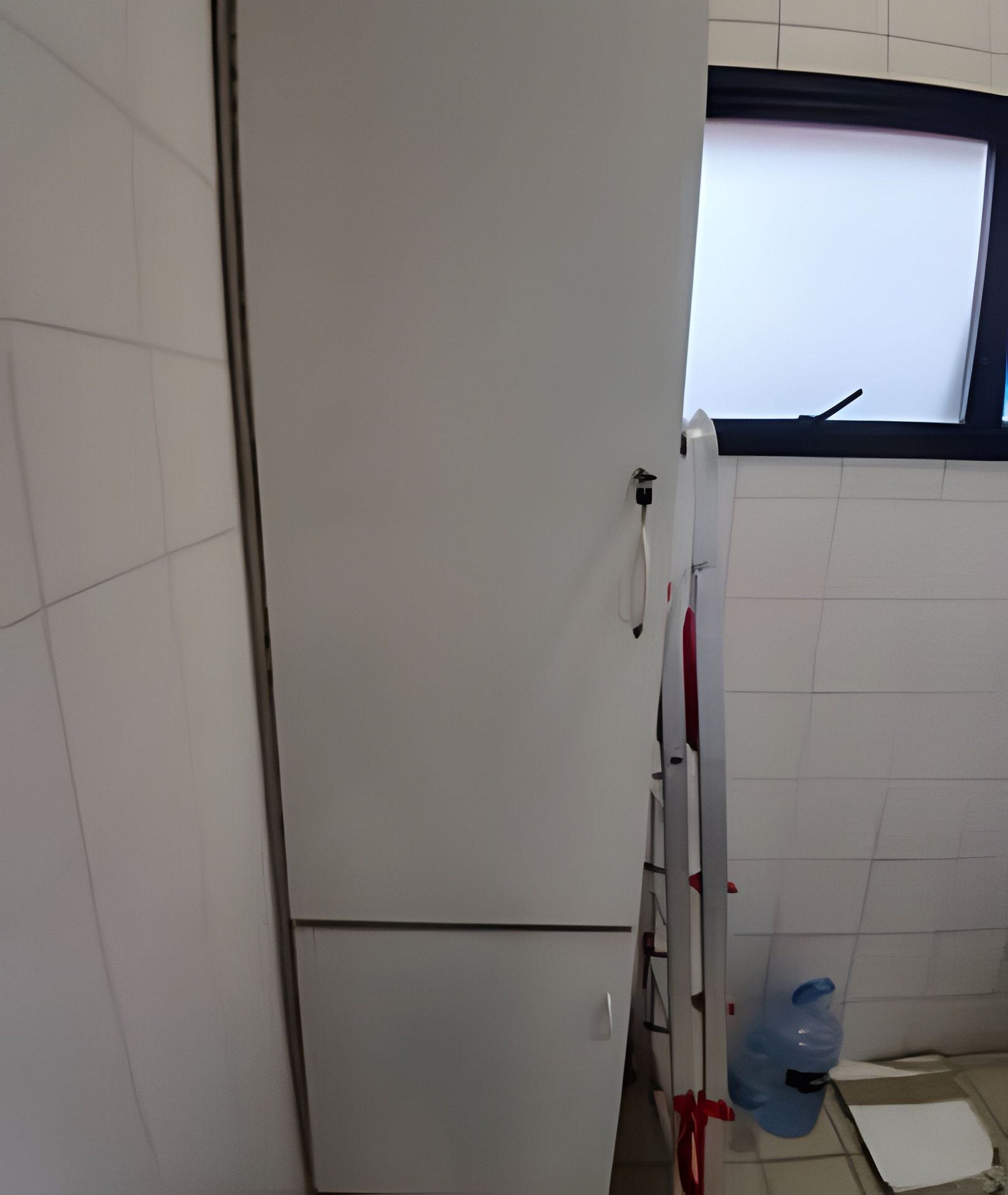 A bathroom with white tiled walls, a window with a black frame, a white towel hanging on a rack, and a blue water bottle on the floor.