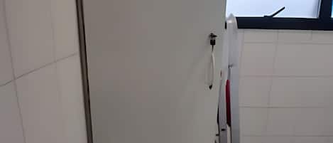 Un baño con paredes de azulejos blancos, una ventana con marco negro, una toalla blanca colgada en un perchero y una botella de agua azul en el suelo.