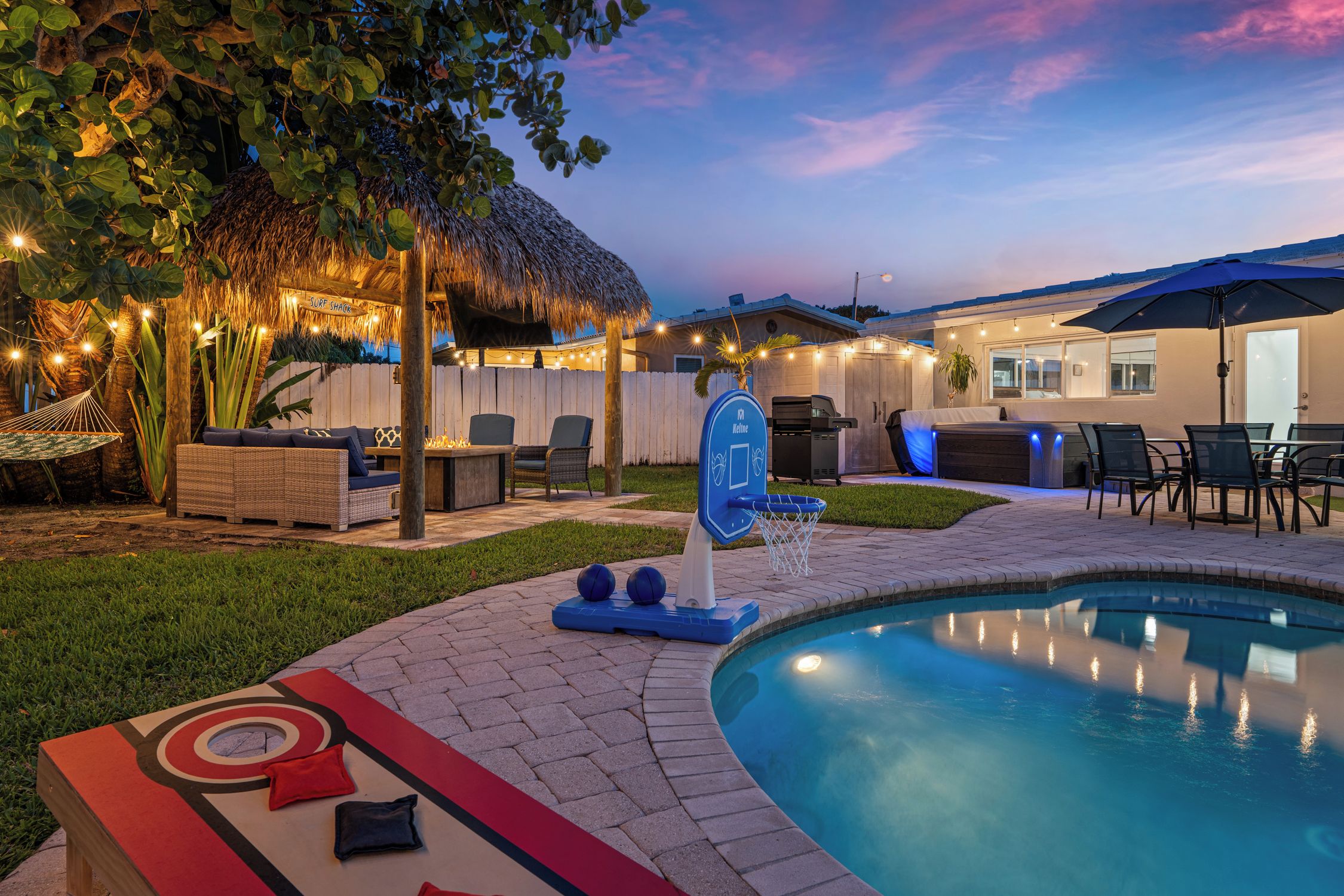 A pool area with a thatched roof, outdoor seating, and a basketball hoop.