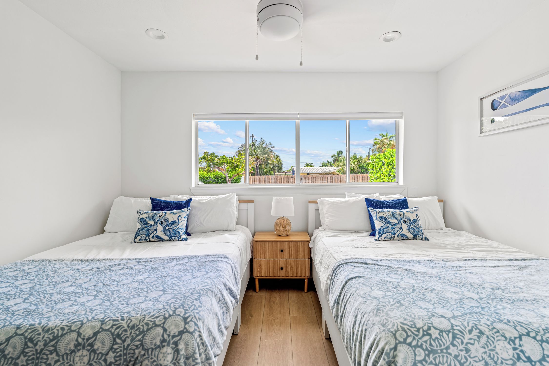 A bedroom with two beds, a window with a view of greenery, and a nightstand with a lamp.