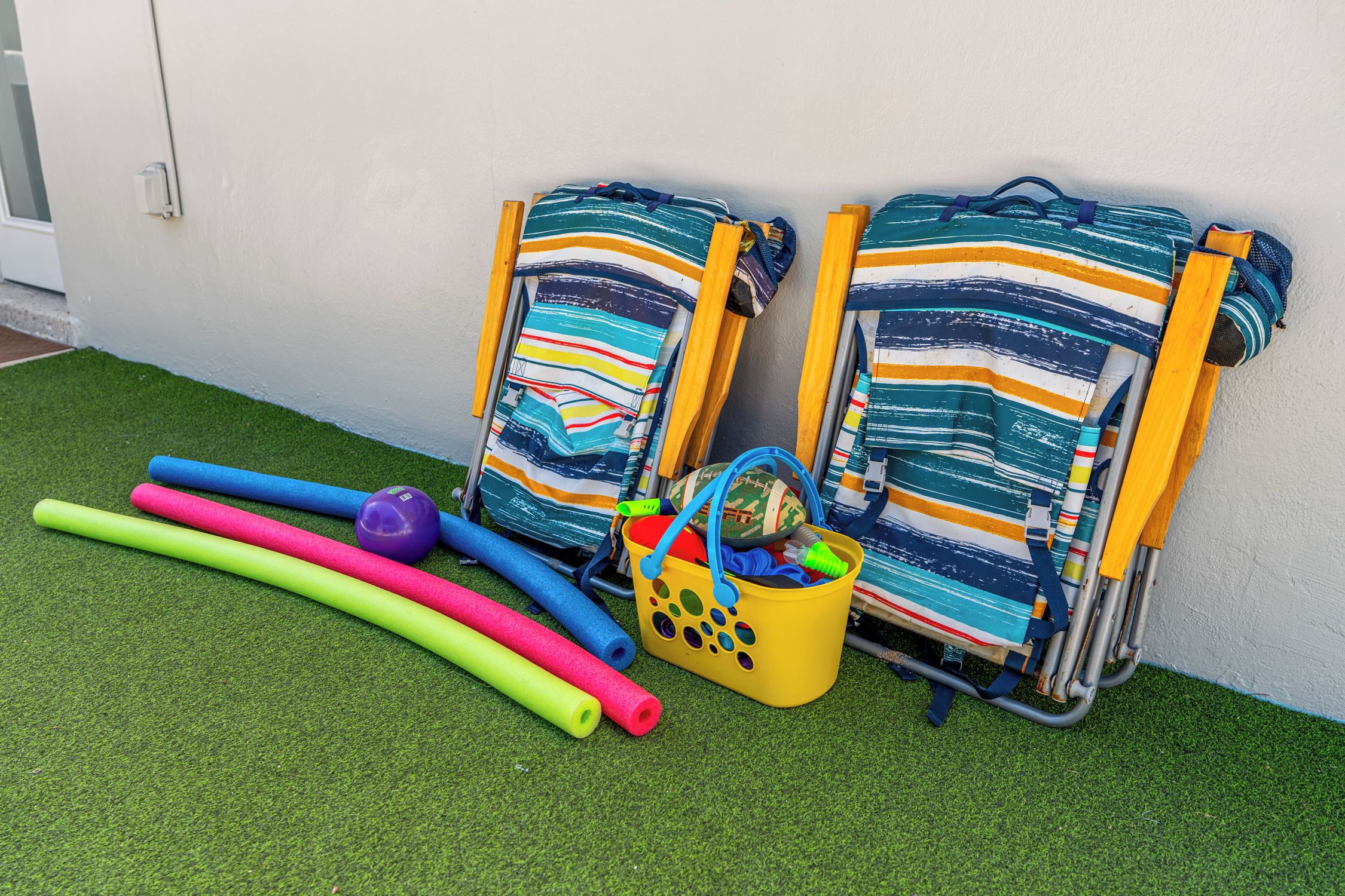 Two beach chairs with striped fabric, a yellow bucket with toys, and colorful foam rollers on artificial grass.
