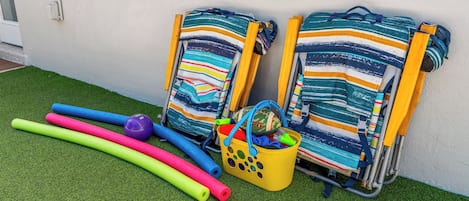 Dos sillas de playa con tela a rayas, un cubo amarillo con juguetes y rodillos de espuma de colores sobre césped artificial.
