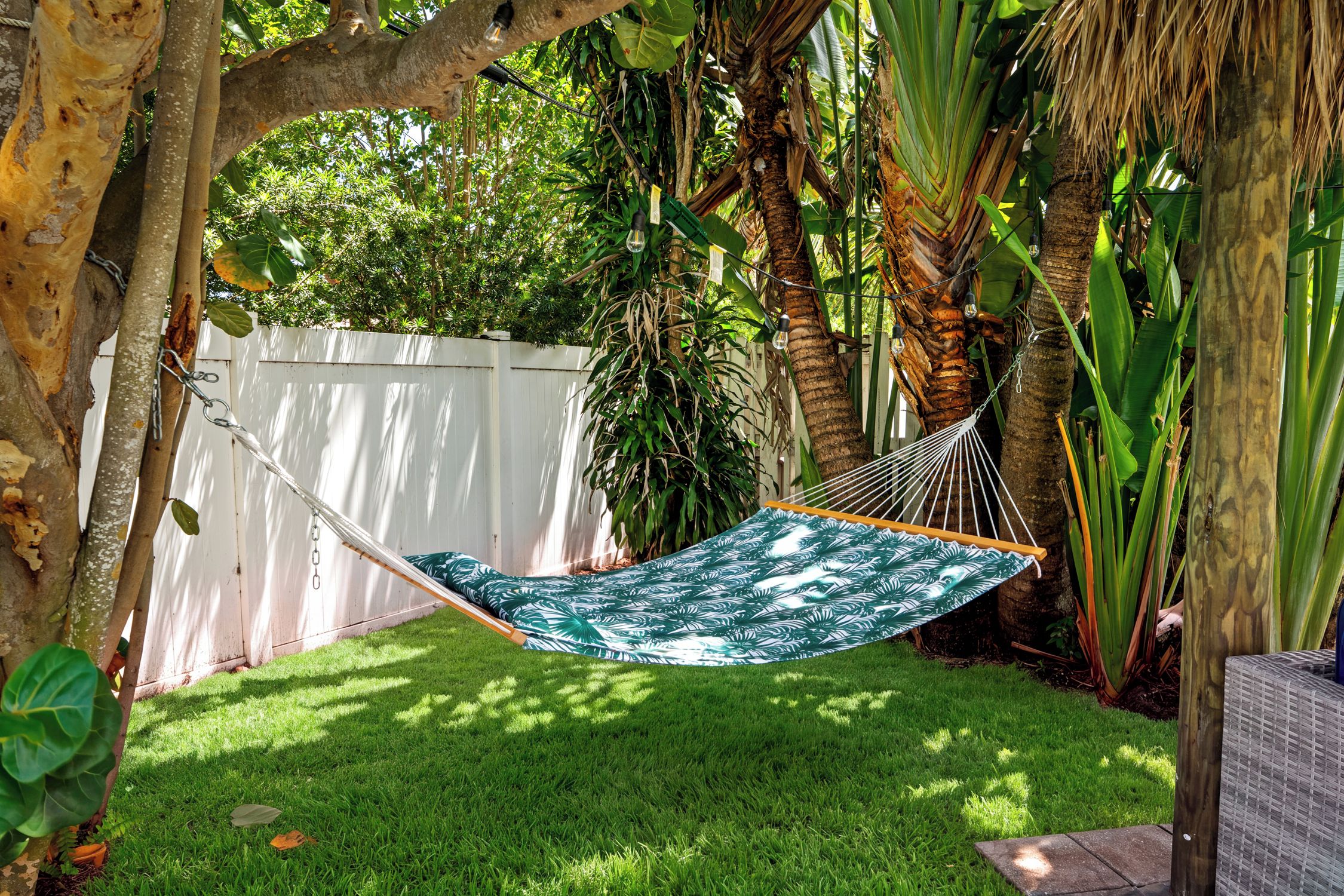 A hammock with a leaf pattern, set in a lush garden with a white fence and various tropical plants.