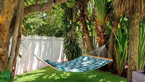 Eine Hängematte mit Blättermuster, platziert in einem üppigen Garten mit weißem Zaun und verschiedenen tropischen Pflanzen.
