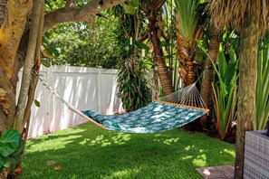 Eine Hängematte mit Blättermuster, platziert in einem üppigen Garten mit weißem Zaun und verschiedenen tropischen Pflanzen.