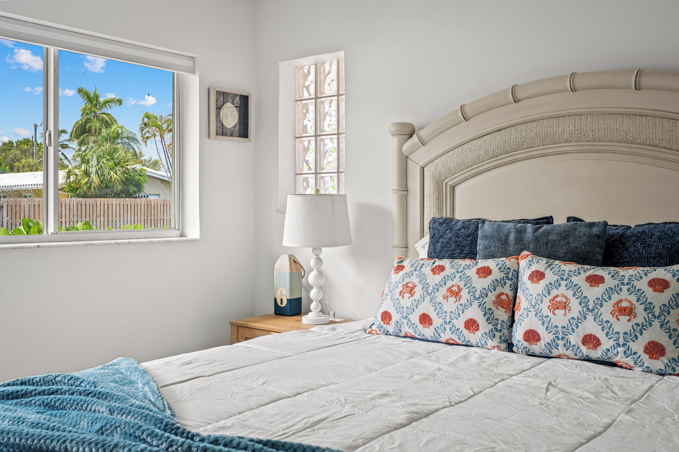 A bedroom with a bed, a nightstand, a window with a view of palm trees, and a wall-mounted picture.