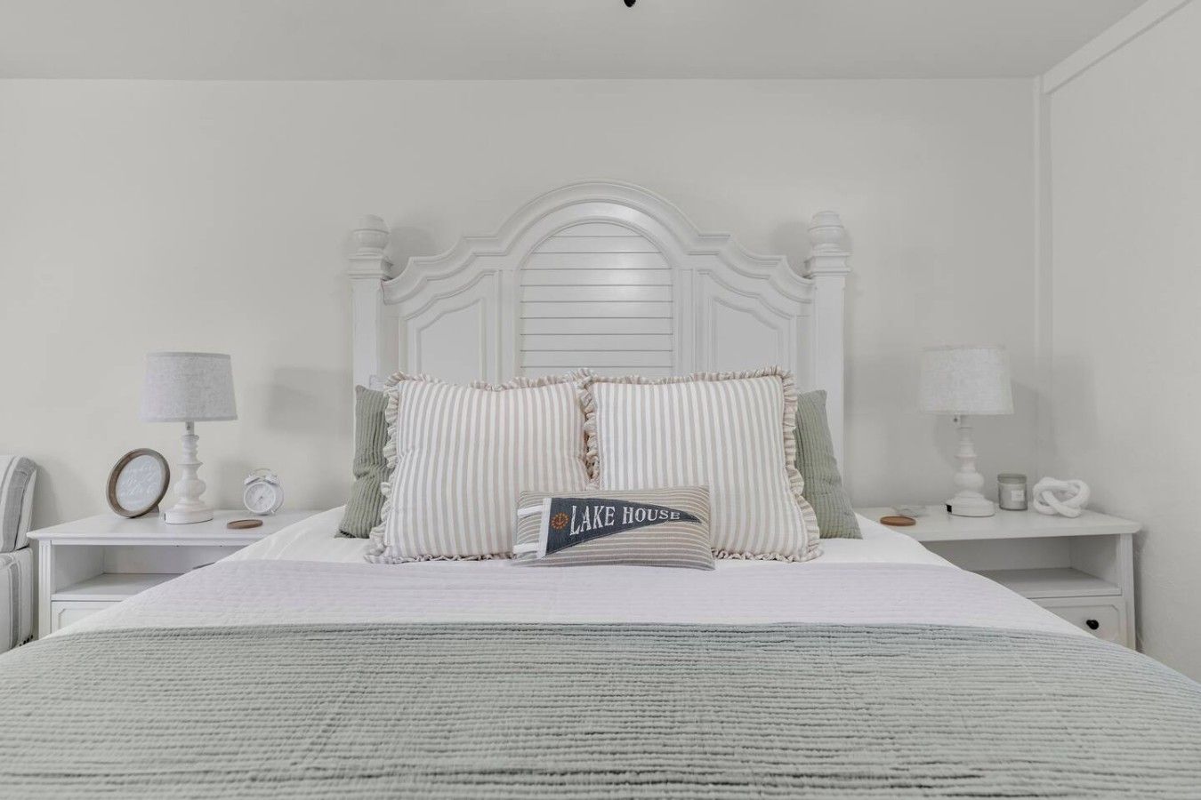 A bedroom with a white bed, two bedside tables, a lamp, a clock, and a pillow with 'LAKE HOUSE' text.