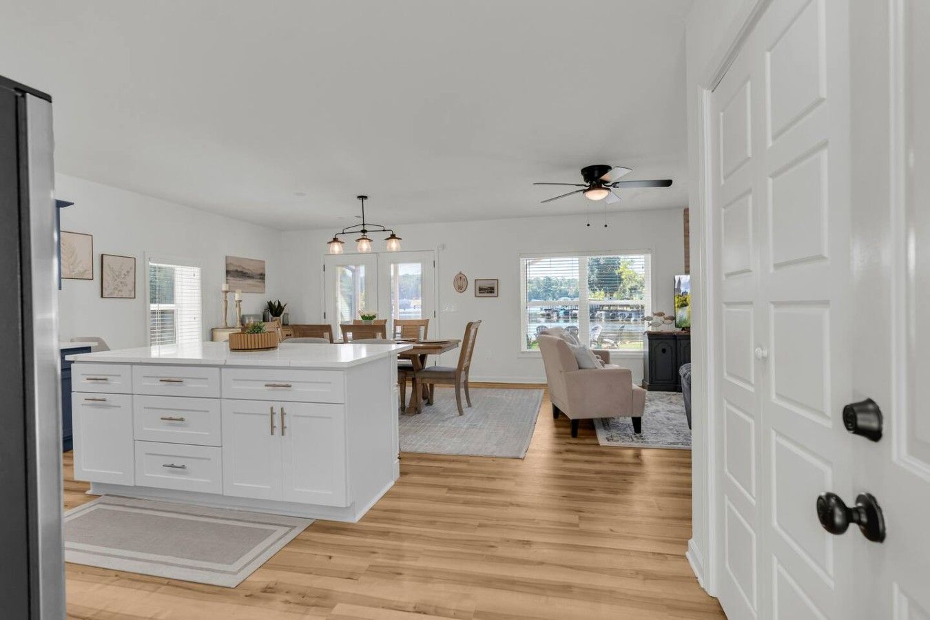 A modern kitchen with white cabinetry, a dining area with wooden chairs, and a view of the outdoors through large windows.