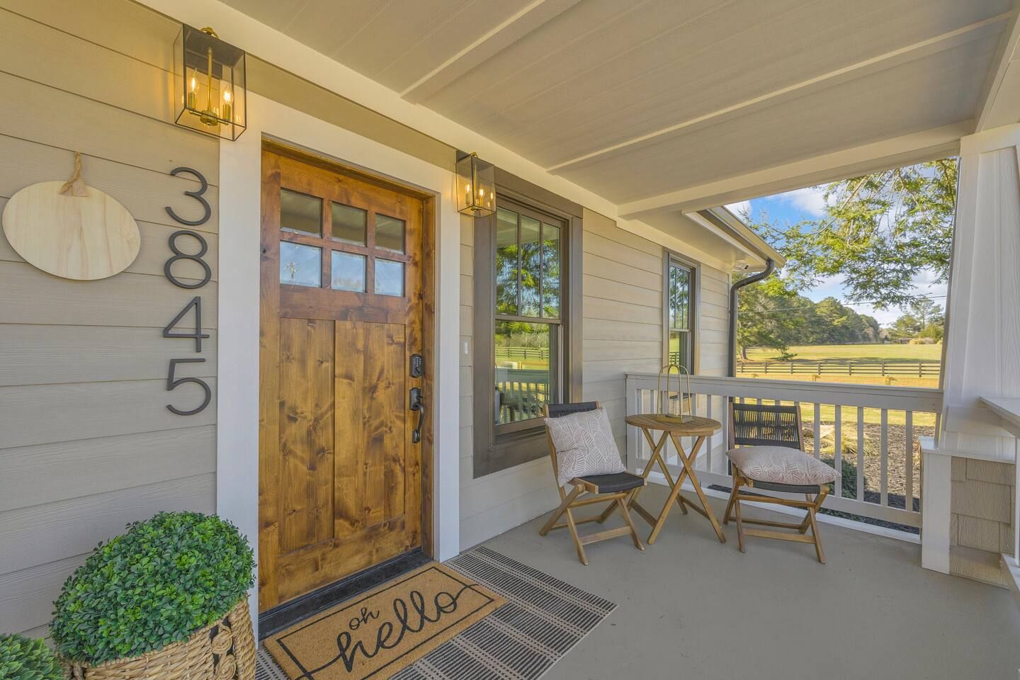 A wooden front door with a number '3845' above it, a porch with two chairs and a small table, and a view of a green landscape through the windows.