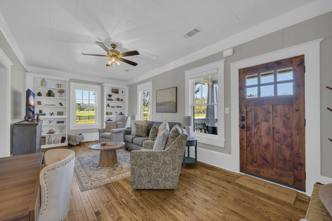 A spacious living room with a ceiling fan, a wooden dining table, and a large window with a view of the outdoors.