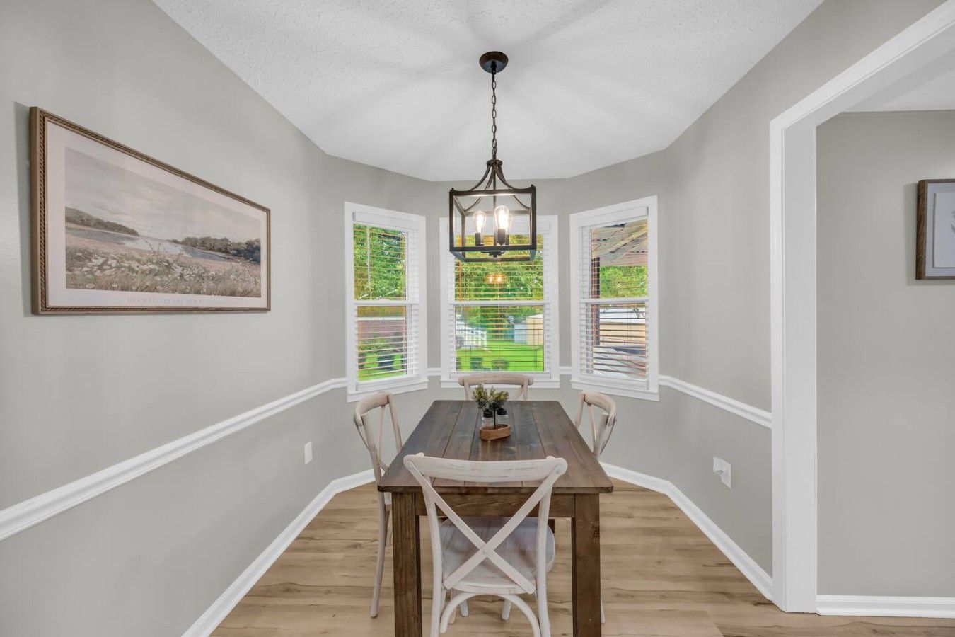 A dining area with a wooden table and chairs, a hanging light fixture, and a view through a window with blinds.