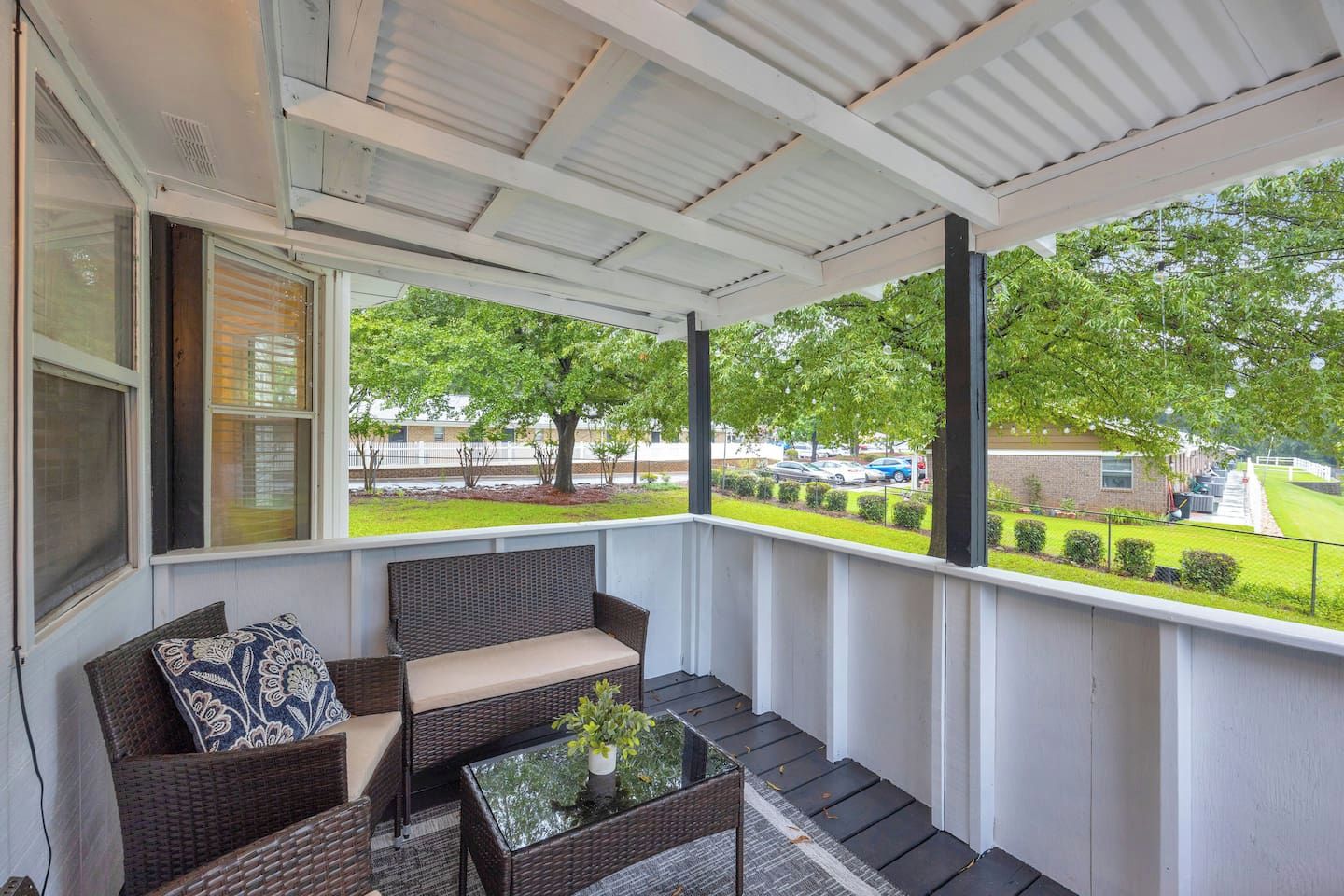 A covered porch with wicker furniture, a glass table, and a view of a parking lot and greenery.