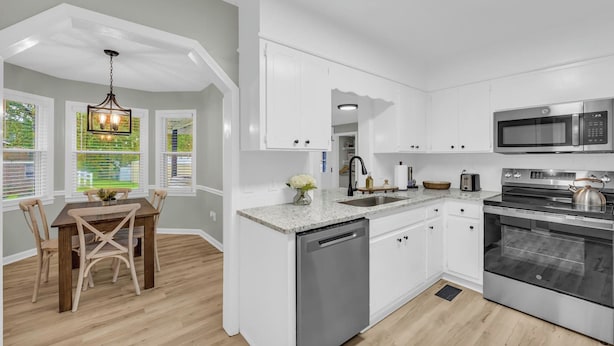 A modern kitchen with white cabinetry, stainless steel appliances, a dining area with a wooden table and chairs, and a view through the window.