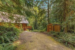 A rustic cabin with a wooden deck and a small shed, surrounded by dense forest.