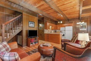 A cozy living room with a wooden staircase, a television, a fireplace, and a kitchen area.