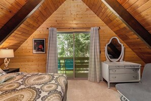 A bedroom with a sloped ceiling, a bed with patterned bedding, a dresser with a mirror, and a view of greenery through a door.