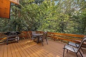 Um deck externo com churrasqueira, mesa e cadeiras, cercado por árvores densas.