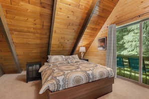 Quarto com teto inclinado, cama com roupa de cama estampada, mesa de cabeceira, abajur e janela com vista para o verde.