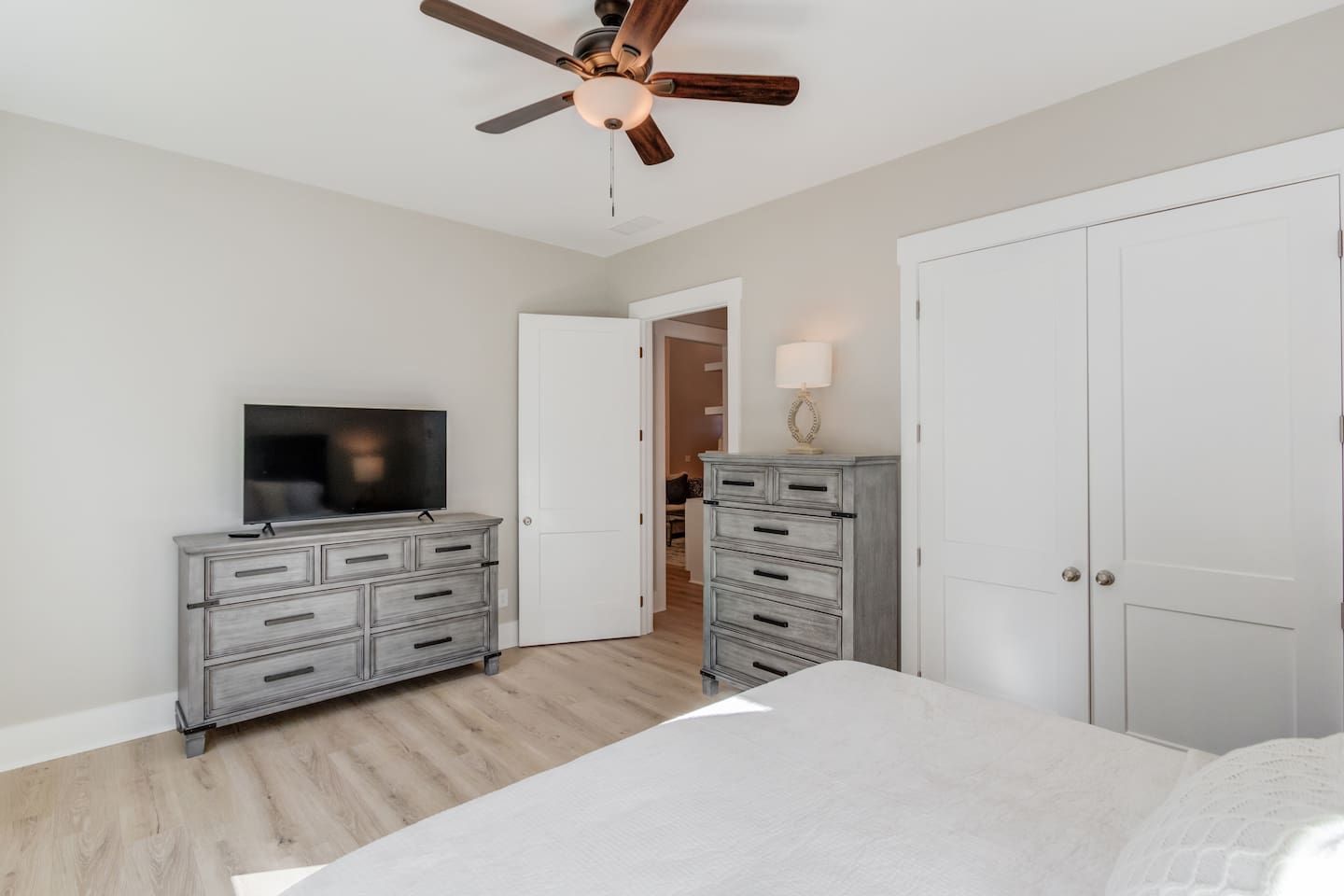 A bedroom with a bed, a dresser, a television, a ceiling fan, and a door leading to another room.