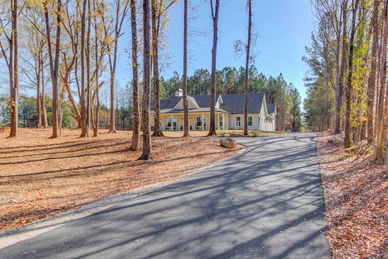 A two-story house with a driveway leading to a gated entrance, surrounded by tall trees and a clear sky.
