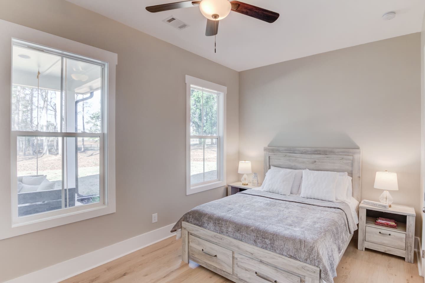 Un dormitorio con cama, dos mesitas de noche, ventilador de techo y ventanas amplias.