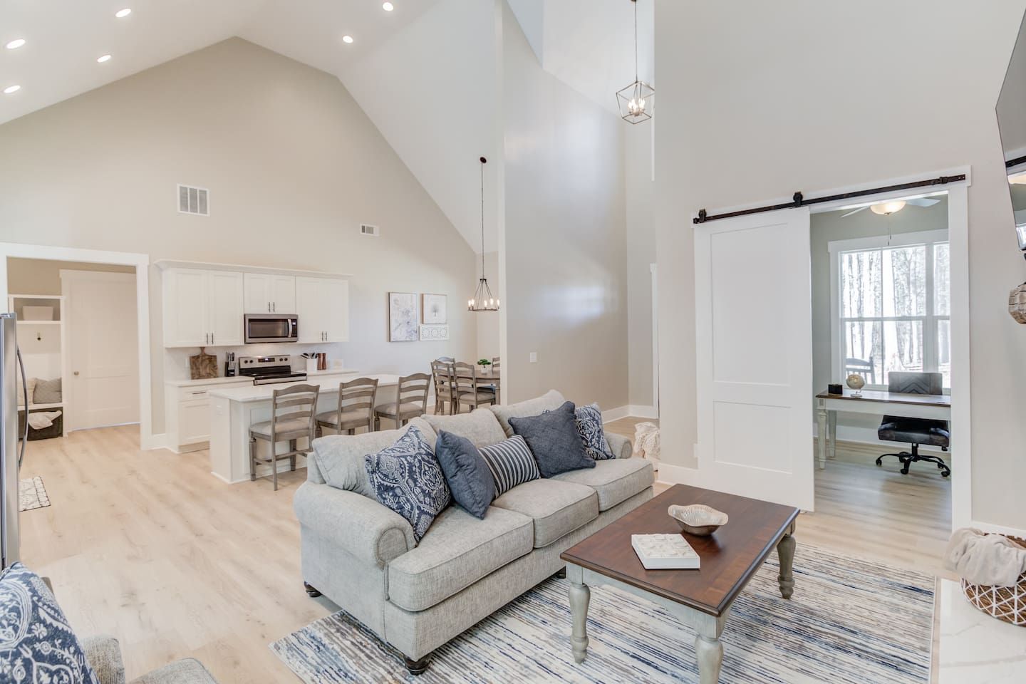 A spacious living area with a high ceiling, a kitchen with white cabinetry, a dining area with wooden chairs, and a large window providing natural light.