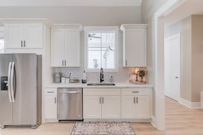 A modern kitchen with white cabinetry, a stainless steel refrigerator, a dishwasher, and a window above the sink.