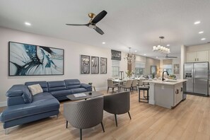 A modern living room with a blue sofa, a dining area with a glass table, and a kitchen with a stainless steel refrigerator. - 4BR Near Disney - Private Heated Pool & Spa (Kissimmee)