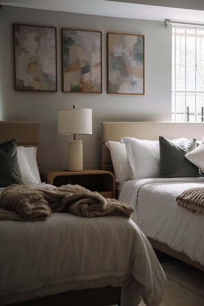 A hotel room with two beds, a nightstand, a lamp, and abstract wall art. - Elegant & Cozy MetLife NYC /2 FREE Parking / Gameroom (Jersey City)