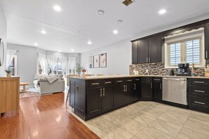 A modern kitchen with dark cabinetry, a stainless steel dishwasher, and a tiled backsplash. - Elegant & Cozy MetLife NYC /2 FREE Parking / Gameroom (Jersey City)