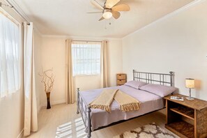 Un dormitorio con cama, mesita de noche con lámpara, ventilador de techo y ventana con cortinas.