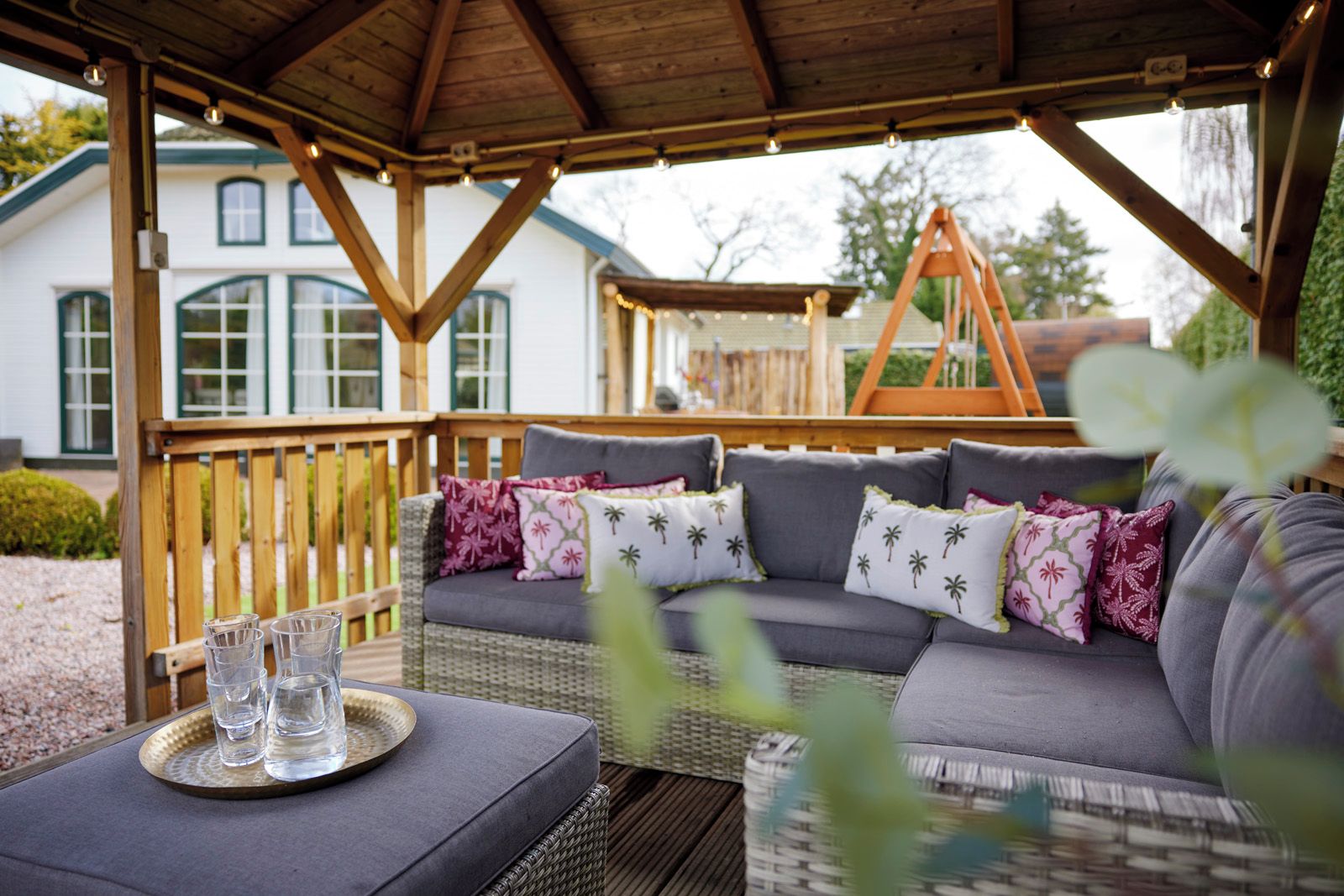 A covered outdoor seating area with a sofa and chairs, a wooden deck, and a house in the background.