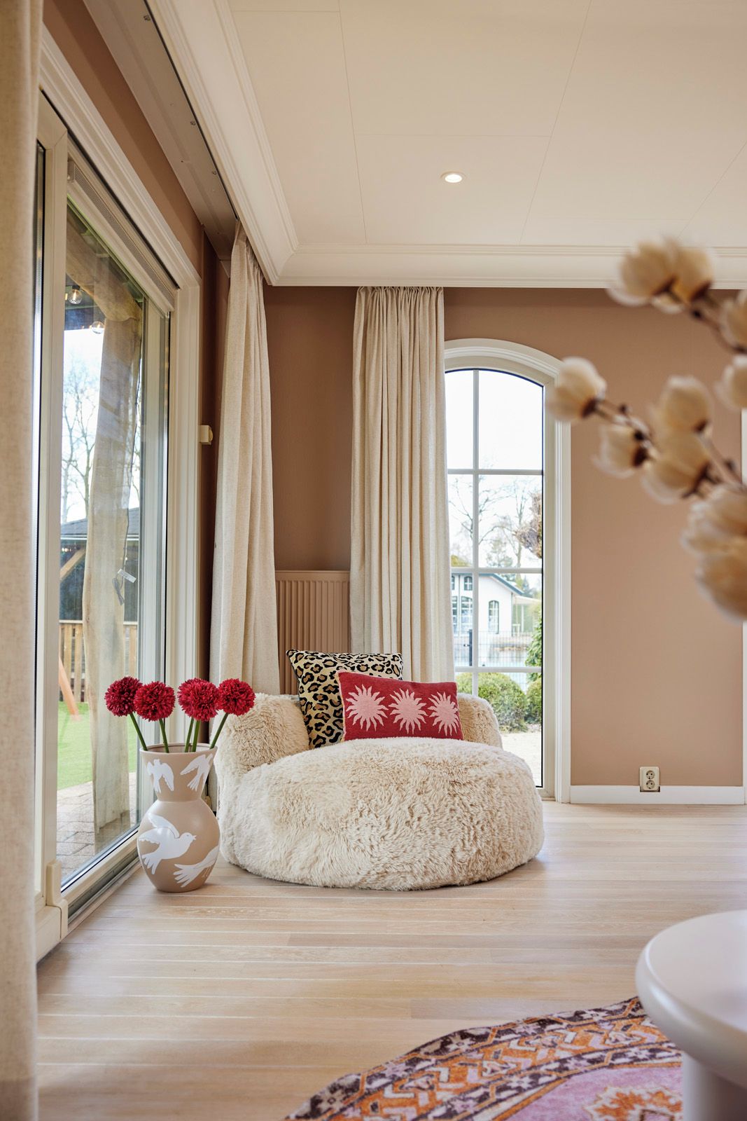 Una habitación con un ventanal grande, un sofá blanco mullido y un jarrón con flores rojas.