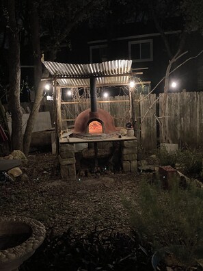 Un four en briques installé à l’extérieur, surmonté d’un toit en métal, entouré d’une clôture en bois, sous un ciel nocturne.
